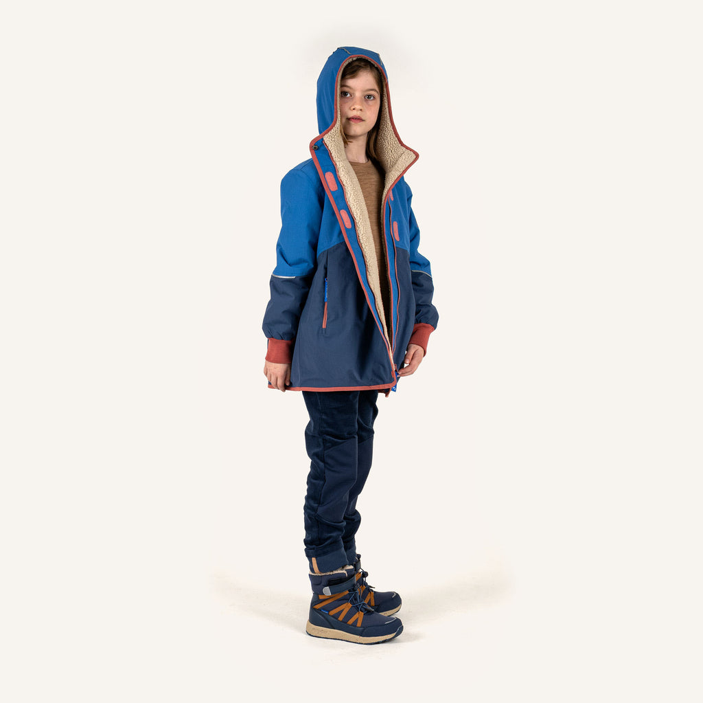 Kinderregenjacke mit Kapuze, blau, dunkle Hose und Skischuhe als Größenbezug